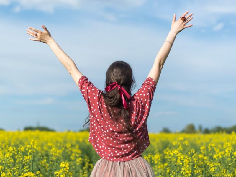 A woman in a polka dot shirt raising her arms in a bright yellow flower field, expressing freedom and joy.