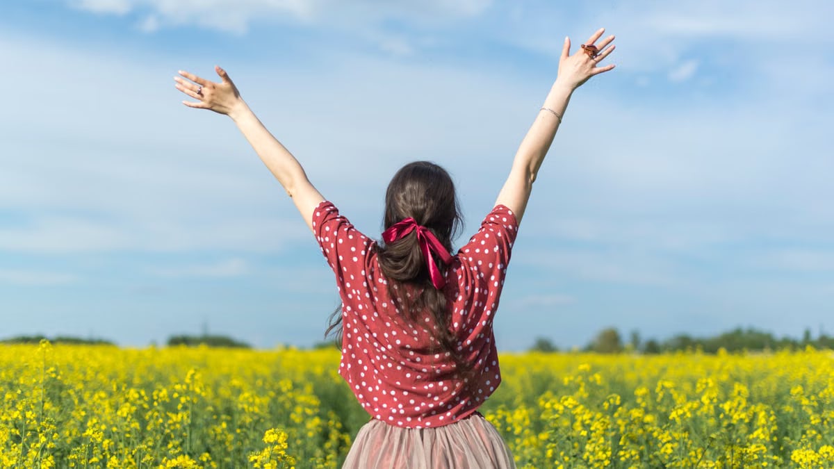 A woman in a polka dot shirt raising her arms in a bright yellow flower field, expressing freedom and joy.